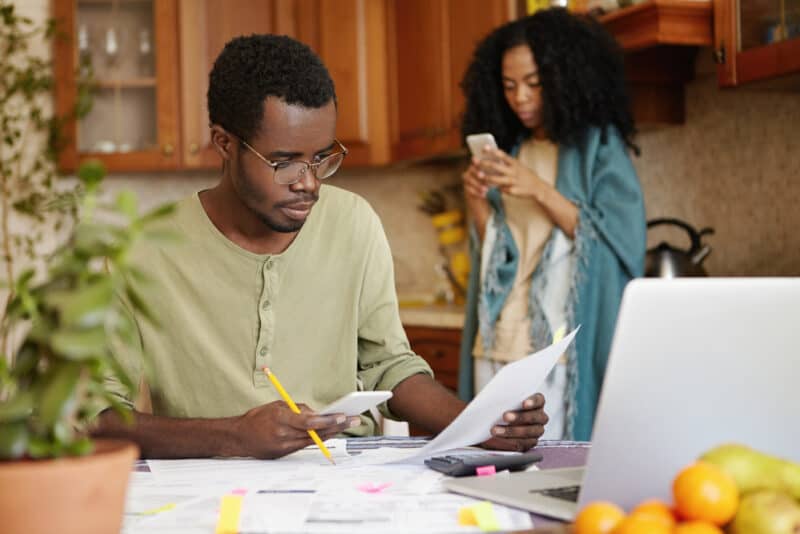 Serious and focused African-American man leading shows paying bills online, using a banking app on his mobile phone. People, paperwork, finances, family budget and economic concept