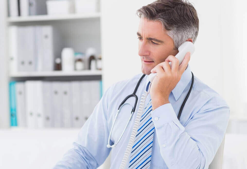 Mature male doctor in his office using a computer while talking on the phone to a patient